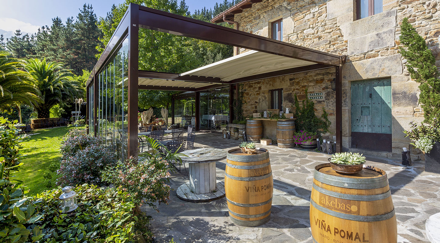 Pergola Balingen mit Glaswänden vor rustikalem Steingebäude